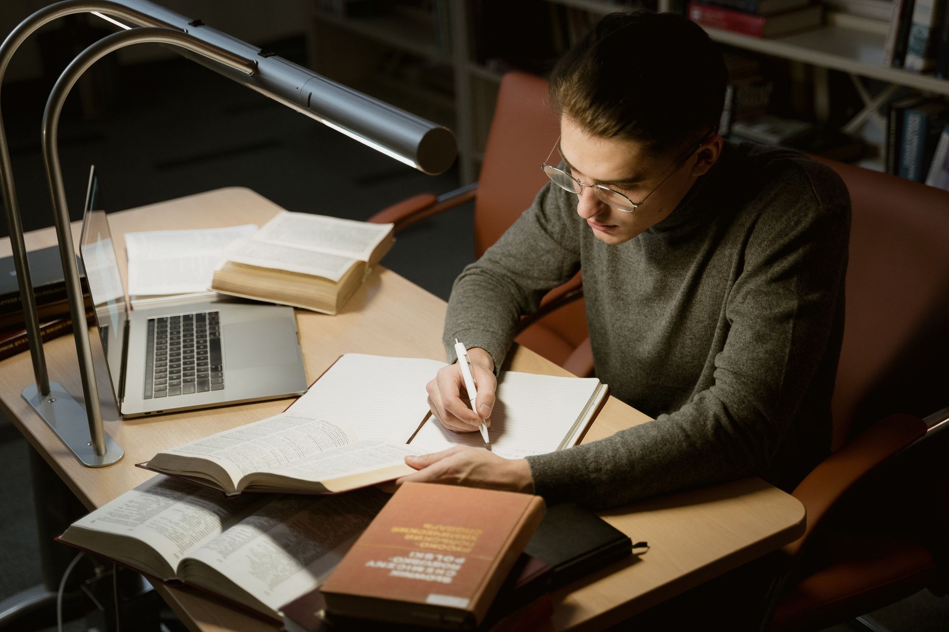 Student przy biurku pisze notatki z książek i laptopa przy lampce